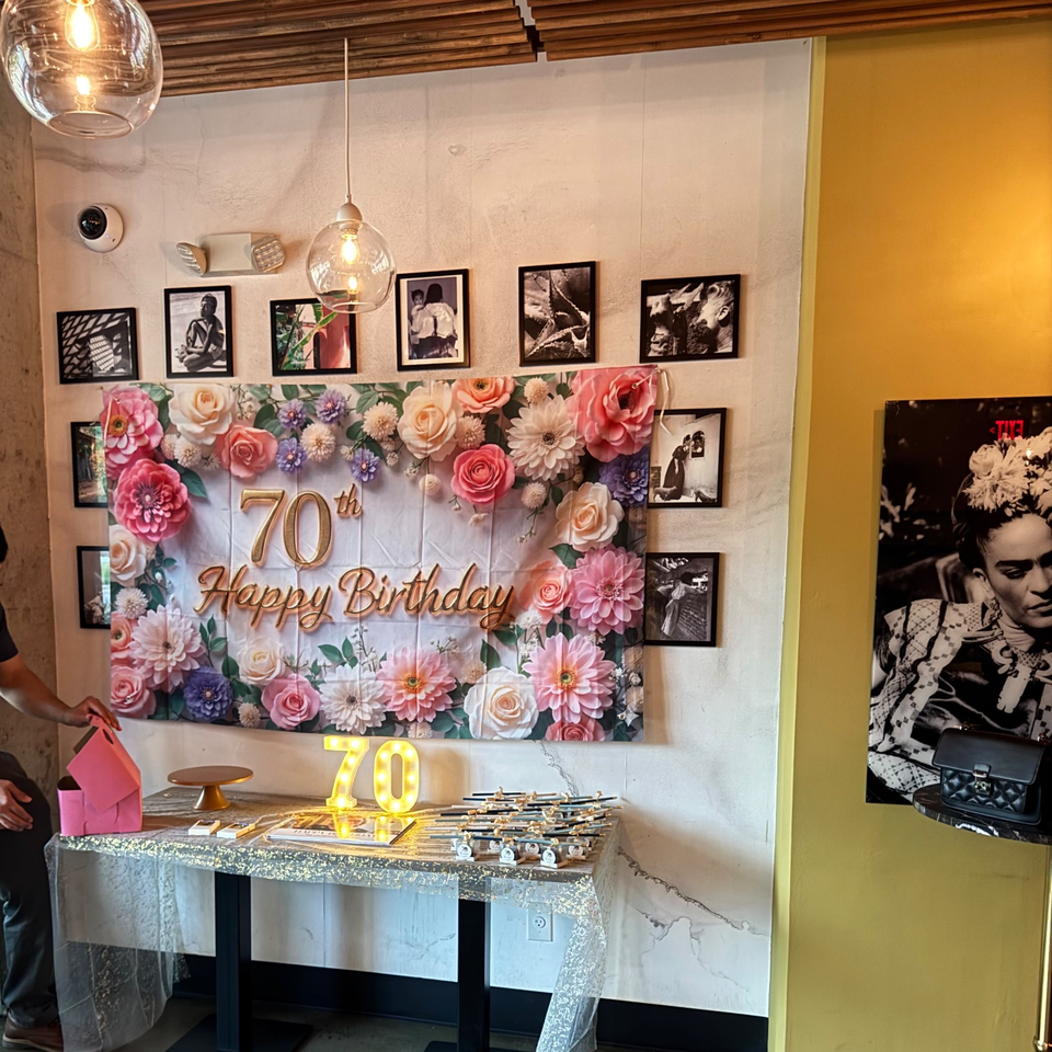 Restaurant celebration display featuring a floral “70th Happy Birthday” backdrop, illuminated number, party favors, framed artwork, and warm pendant lighting.