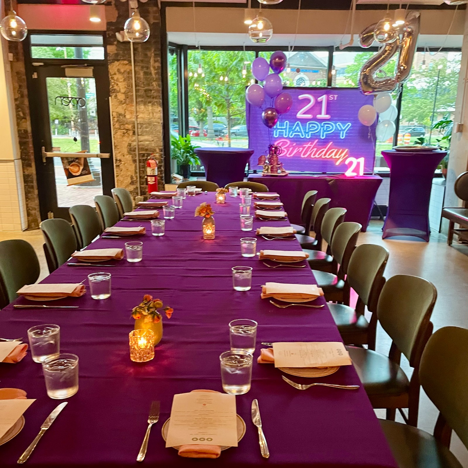 Restaurant dining room decorated for a 21st birthday, with purple tablecloths, place settings, balloons, and a celebratory backdrop.