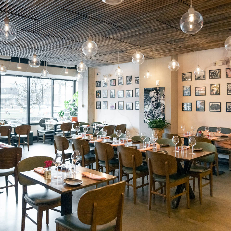 Bright modern restaurant dining room with set tables, wine glasses, pendant lights, framed artwork, and large windows.