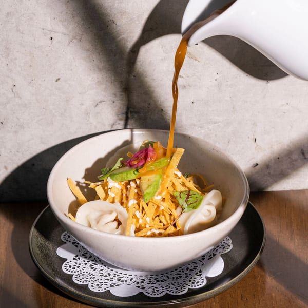 Rich orange broth pours from a white pitcher into a bowl of tortilla soup with dumplings, avocado, and crema.