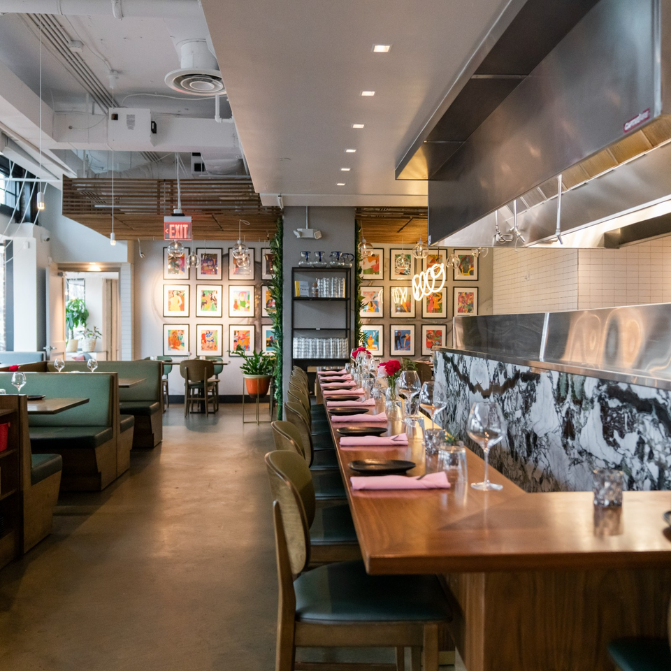 Modern restaurant interior with colorful framed artwork, a marble-topped counter, and tables set with glasses, plates, and pink napkins.
